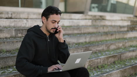 Young-Arabic-handsome-man-with-dark-curly-hair-and-beard-in-black-hoodie-sitting-on-stairs-outside