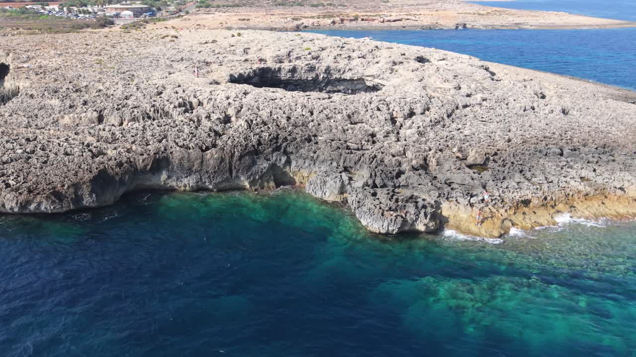 A rocky coastline with visible sea caves along the cliffside, surrounded by deep blue and turquoise waters. The natural landscape contrasts sharply with the calm sea, creating a dramatic coastal scene