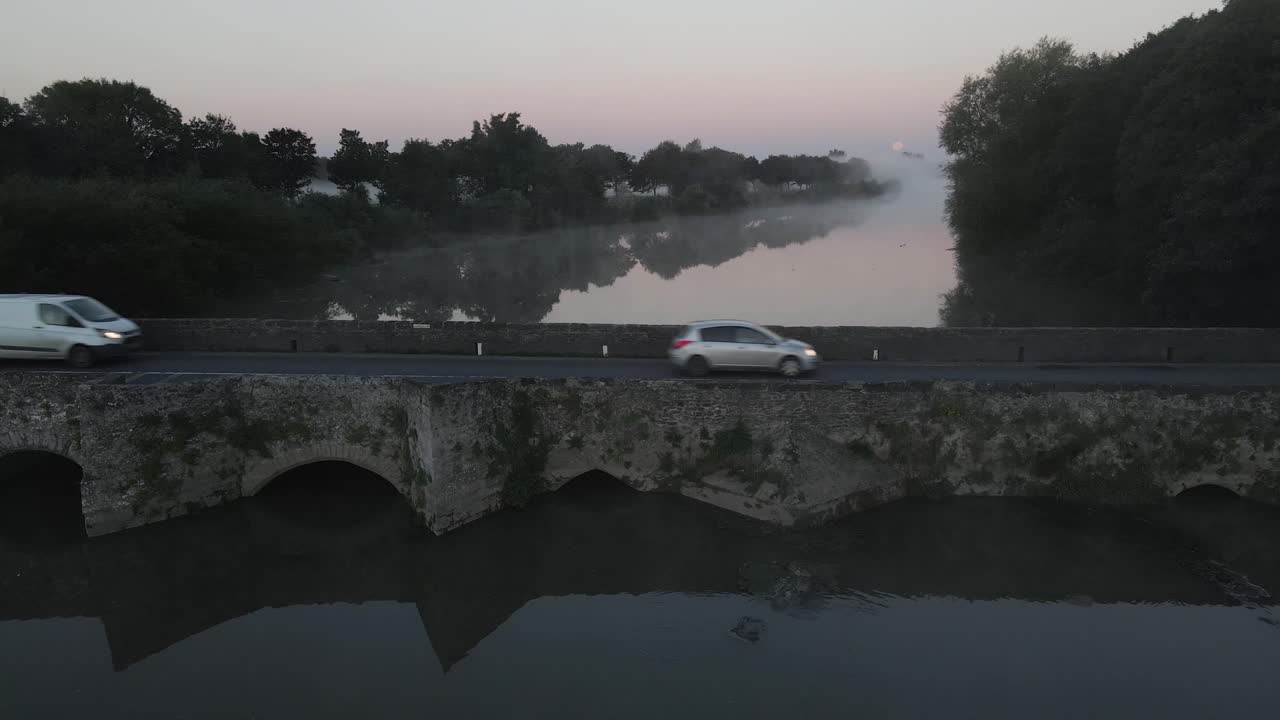 A van and car cross a stone bridge over calm water at dawn in Adare town Ireland