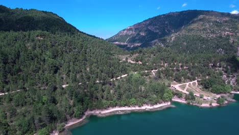 Toma-Aérea-Inclinada-Hacia-Abajo-De-La-Orilla-Del-Lago-Tsivlou-Rodeada-De-Montañas-Con-Pinos-Verdes