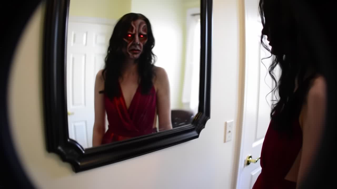 A woman dressed in a bold red outfit stands before a mirror, studying her reflection while adorned with elaborate face paint. The atmosphere hints at a haunting yet captivating vibe.