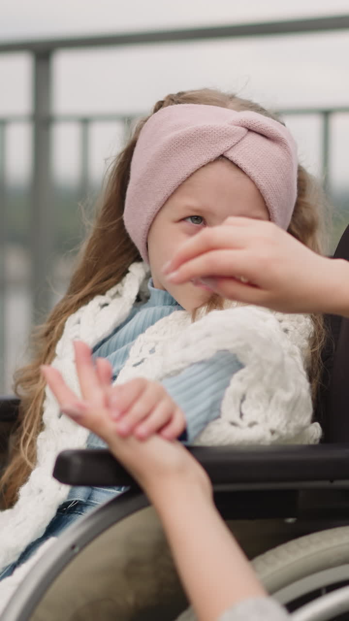 рыжая мать ласкает руку дочери с параличом, выражая любовь. милая девочка с повязкой на голове сидит в инвалидном кресле и смотрит на маму вблизи от забора