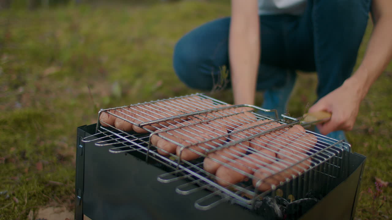 그릴에 소시지 남자는 여름 휴가에서 숲에서 피크닉과 하이킹을 외부에서 요리하고 있습니다.