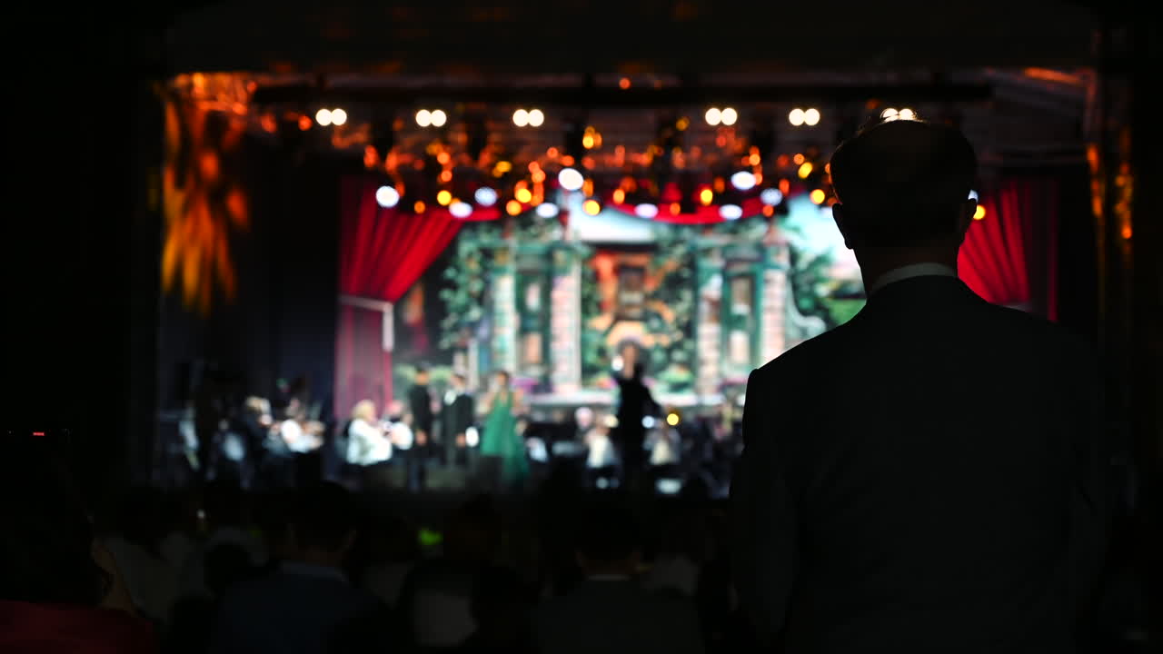 A man stands in the shadows, observing a lively performance filled with musicians and dancers