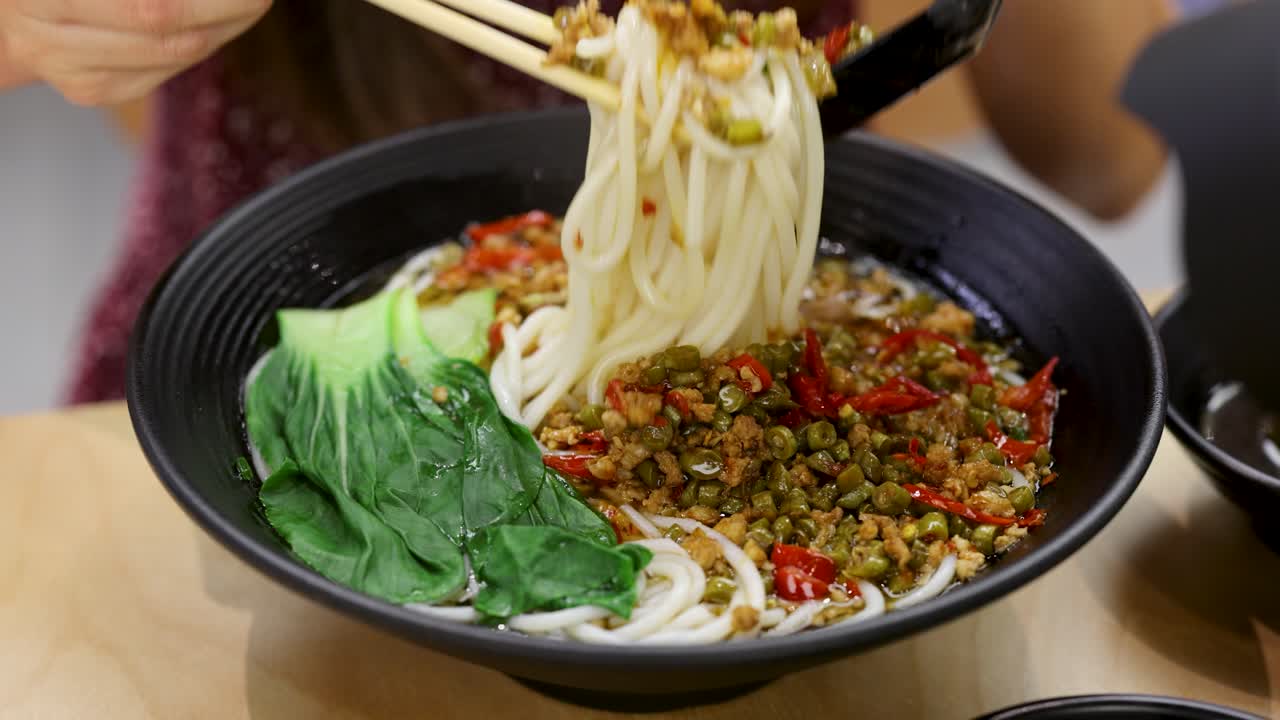 Close-up of woman using chopsticks to lift spicy noodle soup in bright indoor restaurant lighting