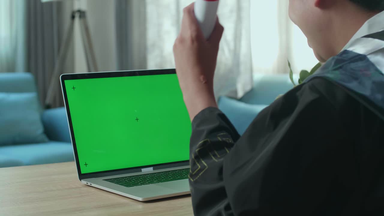 más de la toma del hombro de un hombre asiático celebrando en una computadora portátil con pantalla de maqueta verde durante una llamada de video en línea. graduado masculino con vestido de graduación y gorra sentado en la sala de estar