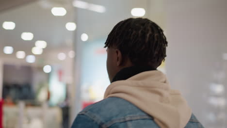 young black man in denim jacket and hoodie walking through bright shopping mall interior looking around store signs and sale displays, rear view capturing shopper exploring retail environment