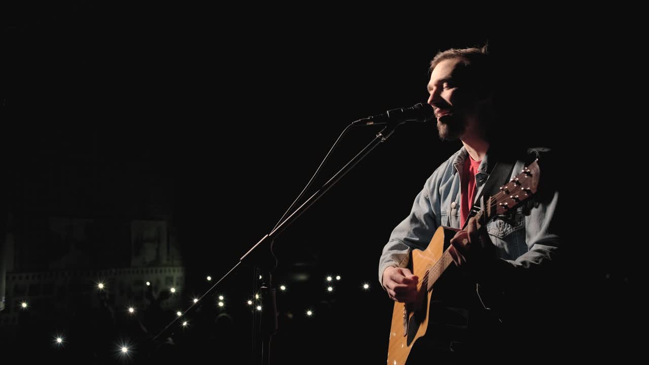 un músico está tocando la guitarra en un gran escenario con miembros de la audiencia sosteniendo linternas en el fondo. el auditorio está lleno de hermosas luces parpadeantes.