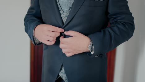 Close up of man adjusting dark blue suit jacket with vest indoors near wooden doorframe on light wall