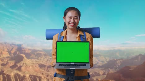 asian female hiker with mountaineering backpack smiling and showing mock up green screen laptop while traveling at the top of mountain