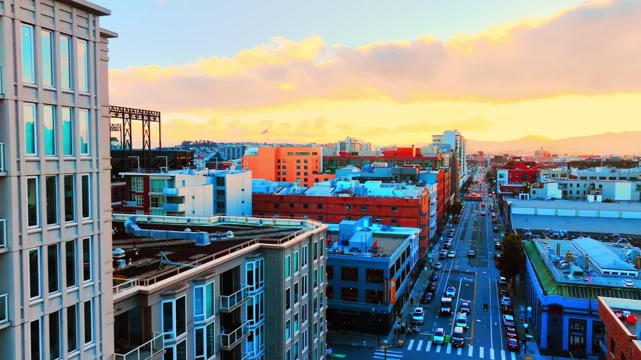 Exteriör vy av ett modernt flerbostadshus i flera våningar med stora glasfönster som reflekterar solnedgångens ljus. Stadens gatunät med fordonstrafik utgör en del av stadsmiljön.