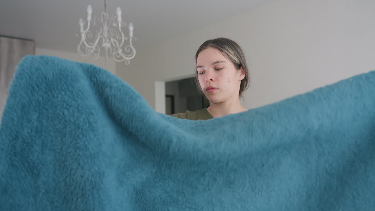 Woman folding fluffy blue blanket indoors, carefully arranging soft fabric in neat folds, natural daylight brightening cozy home setting with chandelier visible in background