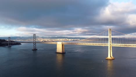 historic san francisco oakland bay bridge - cars travel along the highway, stormy cumulus clouds, calm bay water, sunset reflecting off waves, cinematic landscape tracking down towards water 4k 30fps