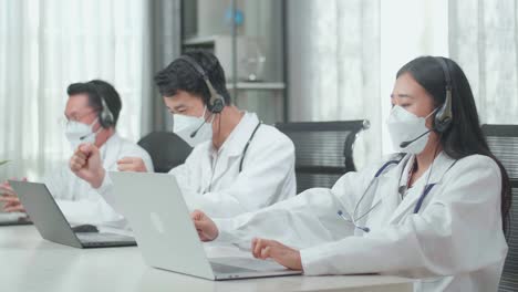 three asian doctors with stethoscopes in headsets and masks working as call center agents sitting on chairs and and being happy due to success working with computers at the office