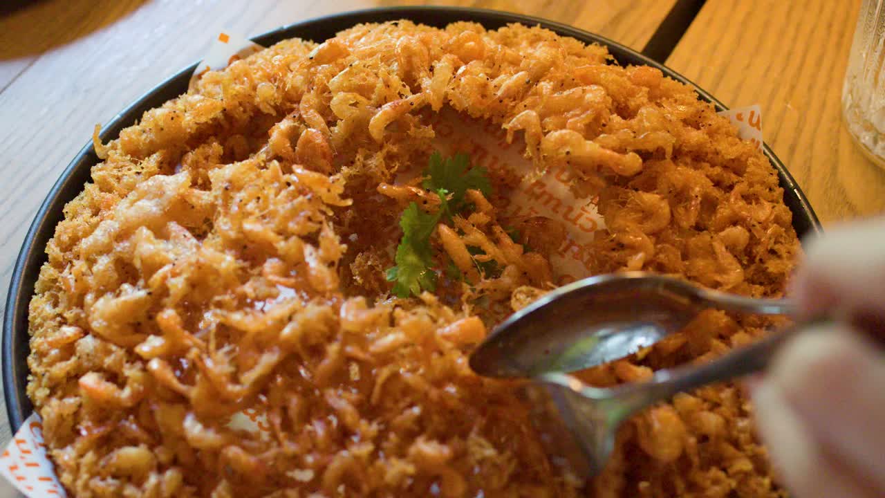 Hands use spoon and fork to serve golden fried shrimp pancake under warm indoor lighting