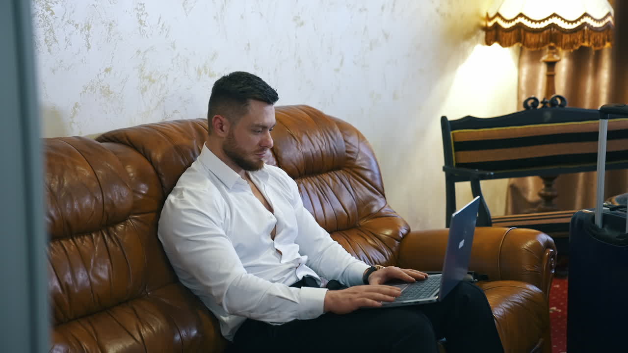Businessman works from the hotel. Young handsome man sitting on a couch and speaking online using laptop. Manager man in business trip in hotel room. Startup business concept.