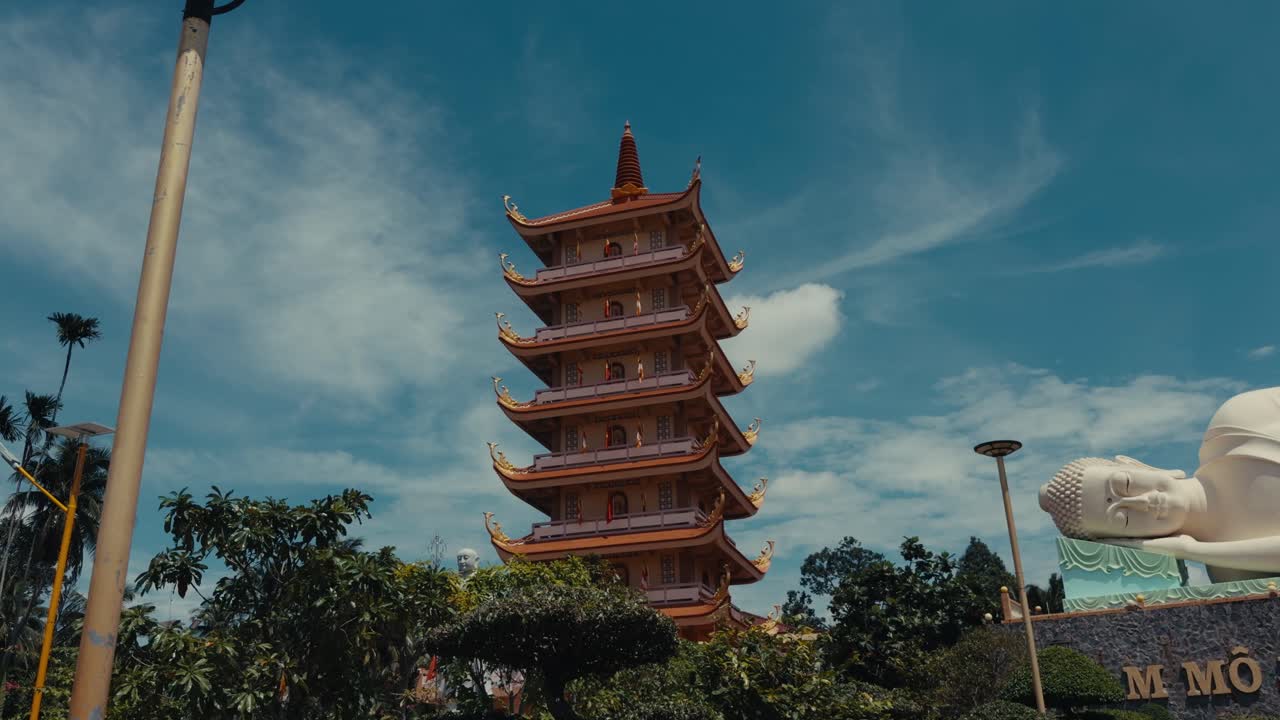 Plano general de una pagoda alta y una gran estatua de Buda reclinado, rodeados de vegetación bajo un cielo azul intenso en Tailandia