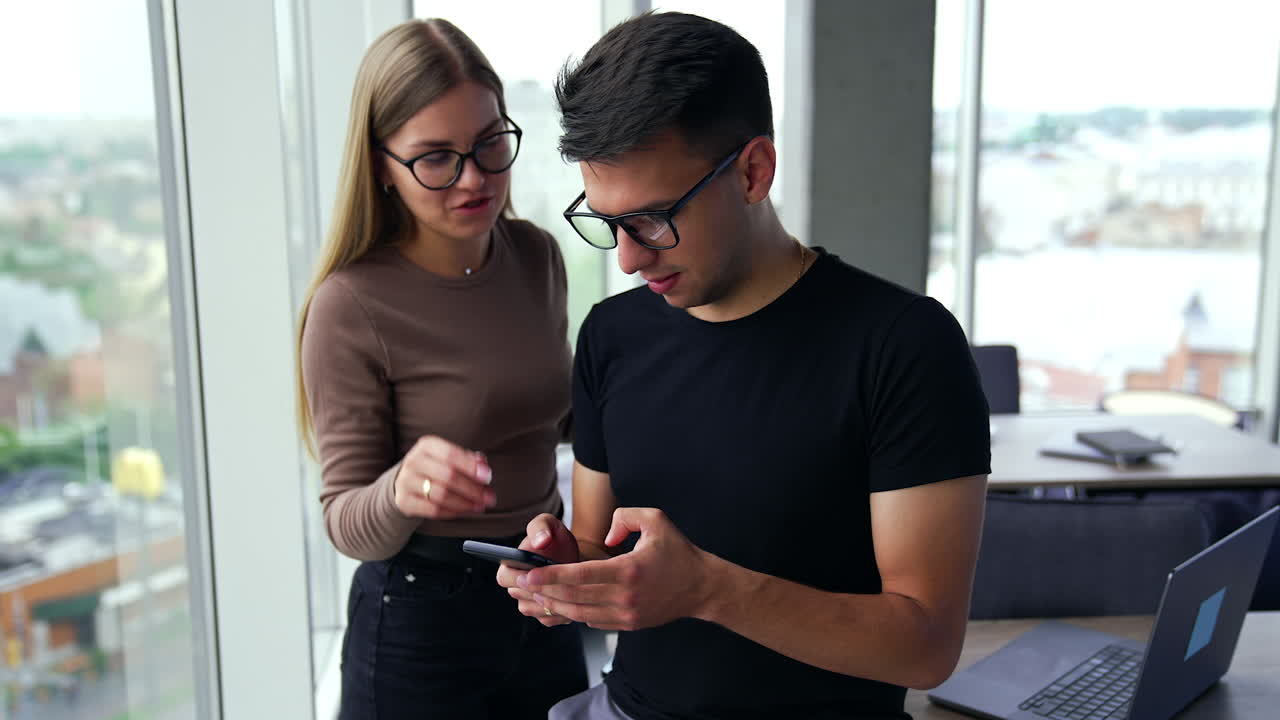 Young people in office discussing some issues during a break from work. Colleagues look at phone in man's hands talking about what they see.
