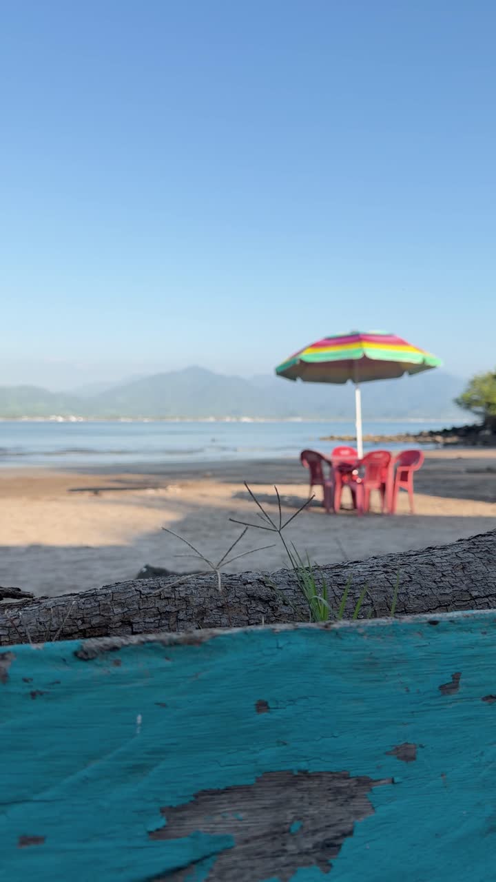Scena verticale e tranquilla di spiaggia sabbiosa con ombrellone colorato e sedie rosse, perfetta per contenuti social di viaggio.