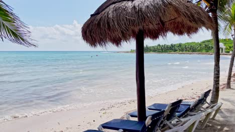 Beautiful-beach-in-Tulum-near-Cancun-Mexico-with-cabanas,-chairs,-small-waves,-palm-trees,-blue-sky-and-some-clouds