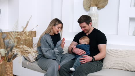 caucasian parents are at home with their baby. child in dad's hands is focused on a bracelet. mom tries to attract her son's attention.