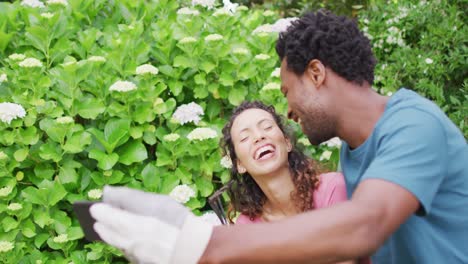 feliz pareja biracial jardinería juntos, tomando selfies con flores