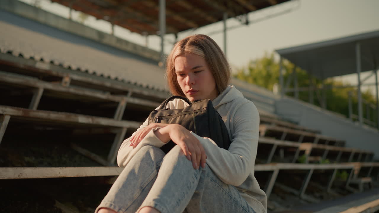 Close-up of lady seated alone on empty stadium bleachers, hugging her bag with a reflective expression, sunlight softly illuminates her as she sits in contemplation