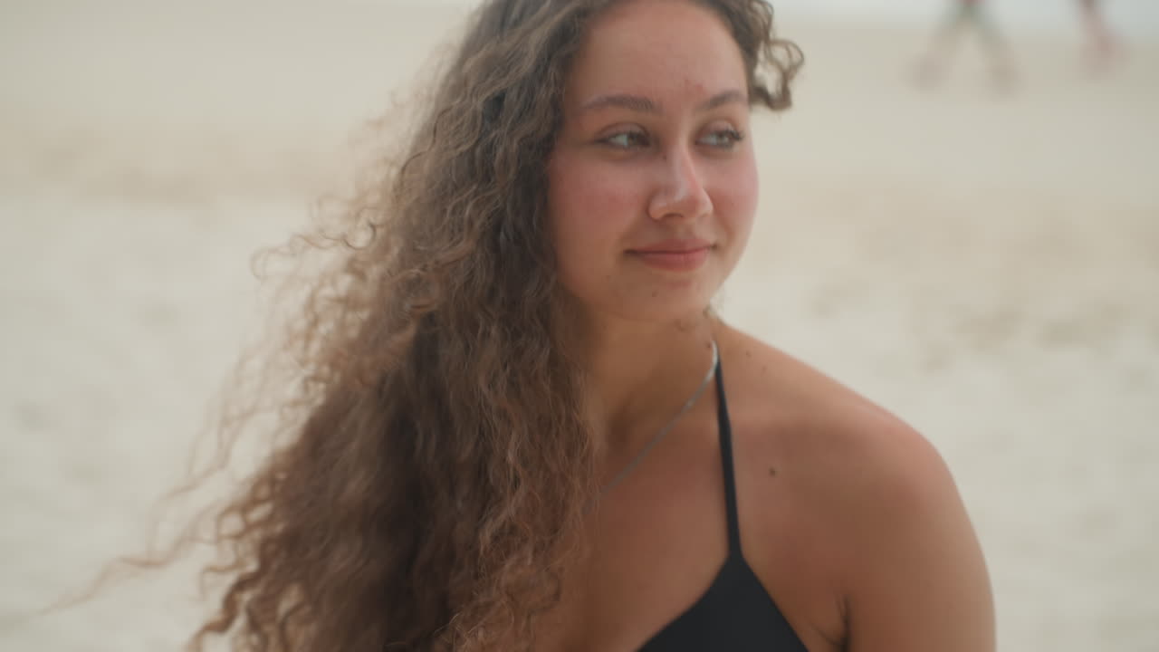 Multietnisk kvinde på østrand i nærbillede, blød solkysset hud og nedslået blik, svagt smil, løse krøller og halskædedetalje, introspektiv stemning, i rollerne som studerende kunstner og mindebevarer