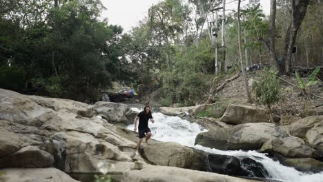 man walking on rocks next to a rushing waterfall in a forest