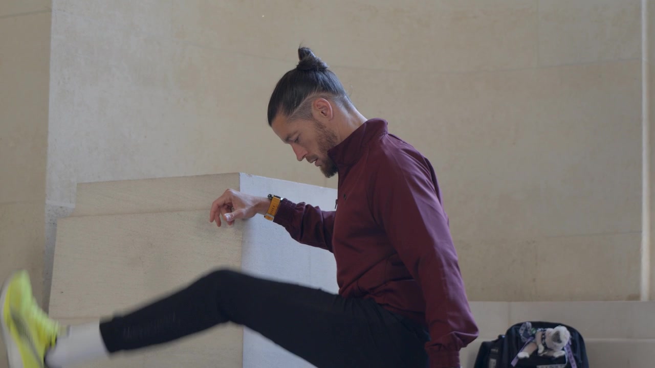 Inquadratura di profilo di un atleta maschio con una giacca bordeaux che slancia la gamba in avanti per il riscaldamento dei muscoli posteriori della coscia, appoggiato a un muro di pietra all'interno.