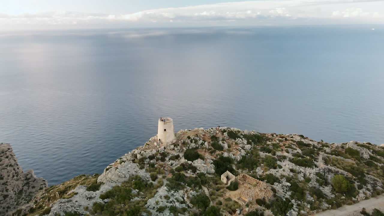 Un drone esegue una dolce orbita a elica attorno alla torre di guardia di Albercutx su un promontorio roccioso, rivelando le rovine circostanti e il vasto e calmo Mar Mediterraneo sotto una tenue luce serale