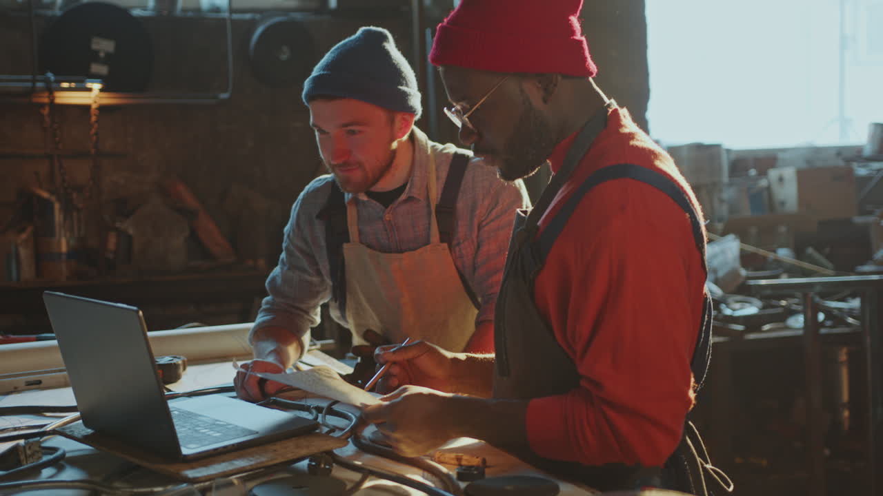 Blacksmith Discussing Metalwork Project on Laptop with Colleague in Workshop