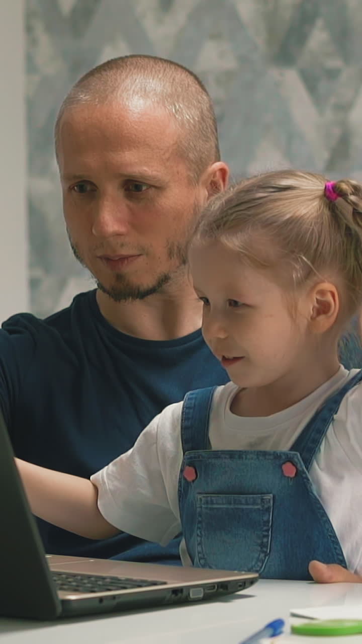 slow motion closeup dad shows her small daughter with blond hair on laptop screen, helps perform school job, they sit at table at home in living room smiling