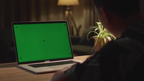 teenage boy sitting at his desk learning online on a laptop with green mock-up screen at home
