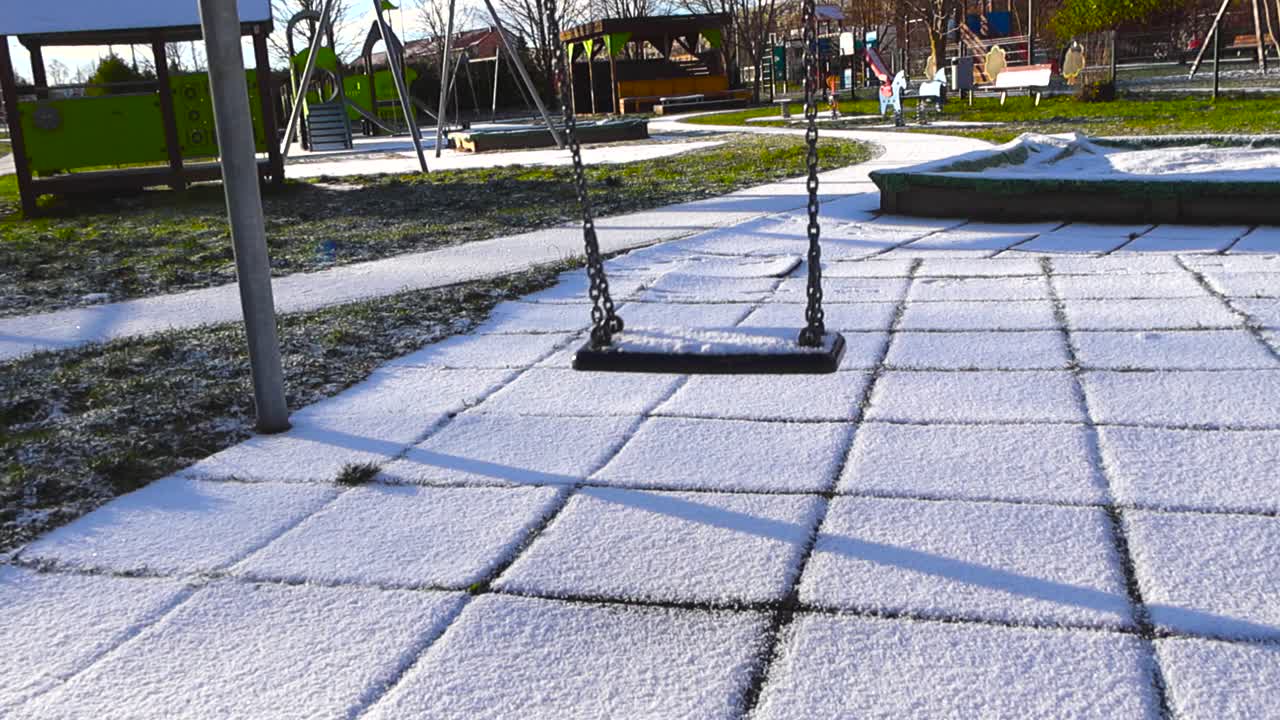Children playground empty swing swining in slow motion back and fourth while covered in fresh white and fluffy first snow during a sunny day, green grass visible between the ice crystals and snow