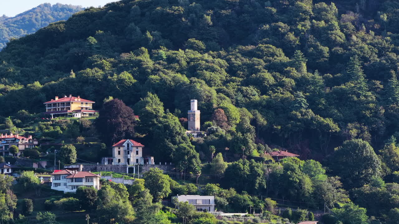 Ripresa aerea cinematografica con drone in volo verso una torre storica e un villaggio collinare circondato da bosco verde vicino a Orta San Giulio e al Lago d'Orta nel nord Italia