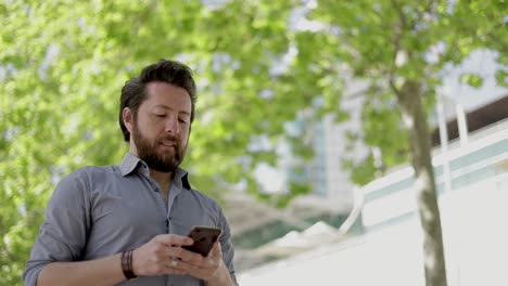 Un-Hombre-Sonriente-De-Mediana-Edad-Enviando-Mensajes-A-Través-De-Un-Teléfono-Inteligente-Al-Aire-Libre