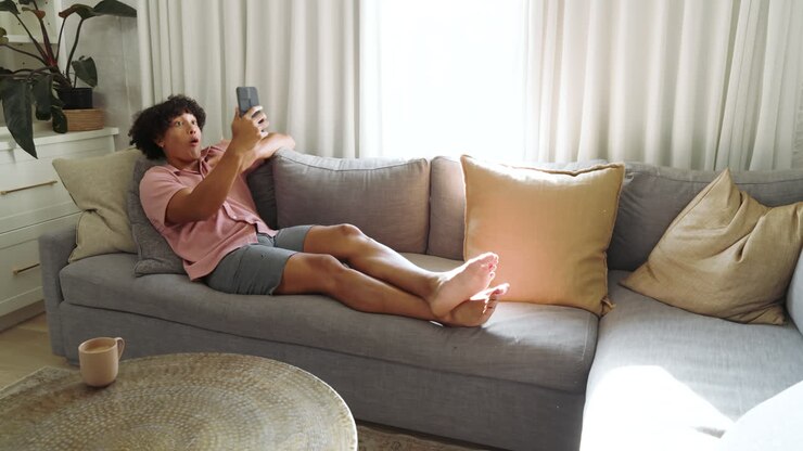Joven relajándose en el sofá, disfrutando de su smartphone en una luminosa sala de estar, en casa, espacio para texto.