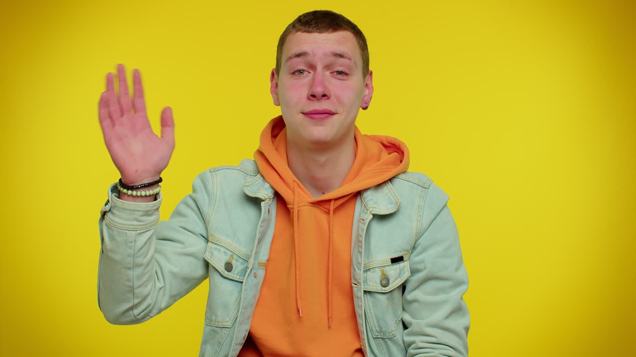 십대 남자가 카메라를 향해 우호적으로 미소 짓고 손을 흔들며 인사하거나 작별 인사를 하고 환영합니다.