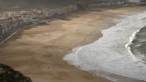 Strand-Von-Praia-Da-Nazaré-Zur-Goldenen-Stunde-Mit-Schatten,-Aufgenommen-Von-Oben-In-4K-Mit-Menschen-In-Der-Ferne,-Die-Am-Strand-Spazieren-Gehen