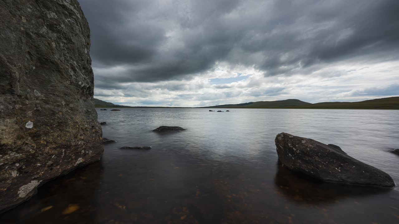 lapso de tiempo de movimiento panorámico del lago con hierba y grandes rocas en primer plano en un oscuro día de verano nublado en el paisaje rural de irlanda