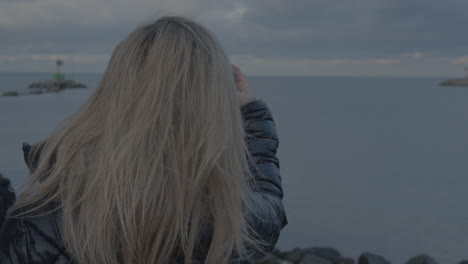 a woman takes a smartphone photo of a landscape while standing on the seashore