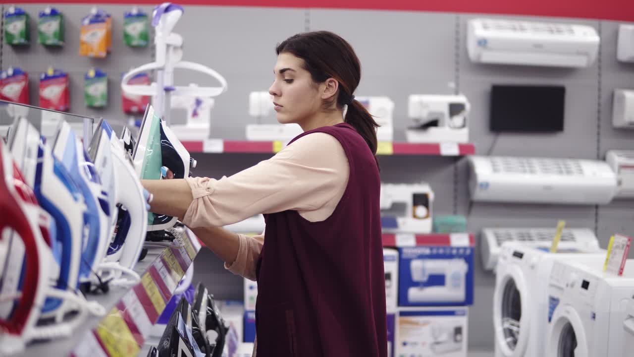 Young brunette girl picks iron from display shelf in hardware store. Looking for household equipment
