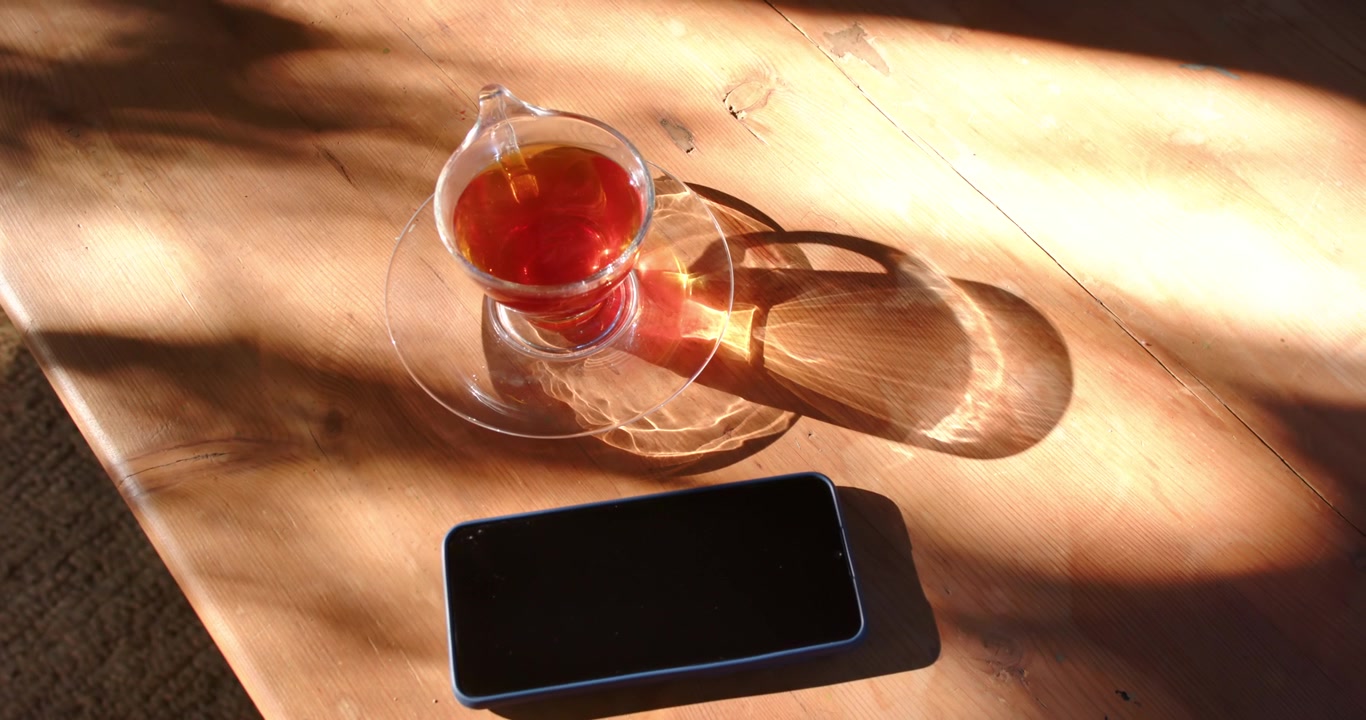 Sunlight sliding across sunlit wooden table is casting ring from teacup and nudging phone clockwise