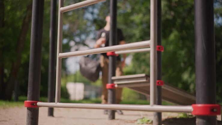 Athlete Examining Outdoor Fitness Equipment, Climber Assessing Exercise Station In Sunlight, Individual Evaluating Fitness Apparatus While Seated On Park Bench Under Shaded Sunlight
