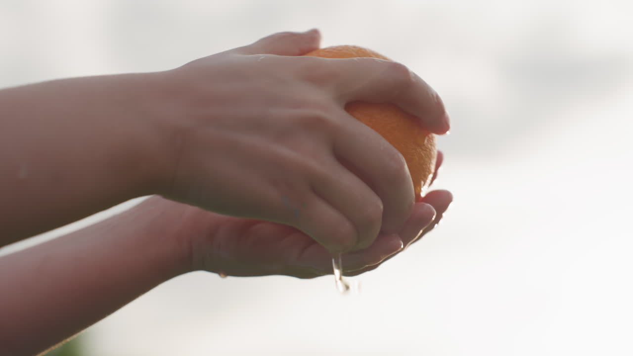 sky view of youngster hands washing orange under sunny sky with backlight, water dripping, healthy outdoor summer snack preparation, closeup on fruit and fingers, bright light creating lens flare