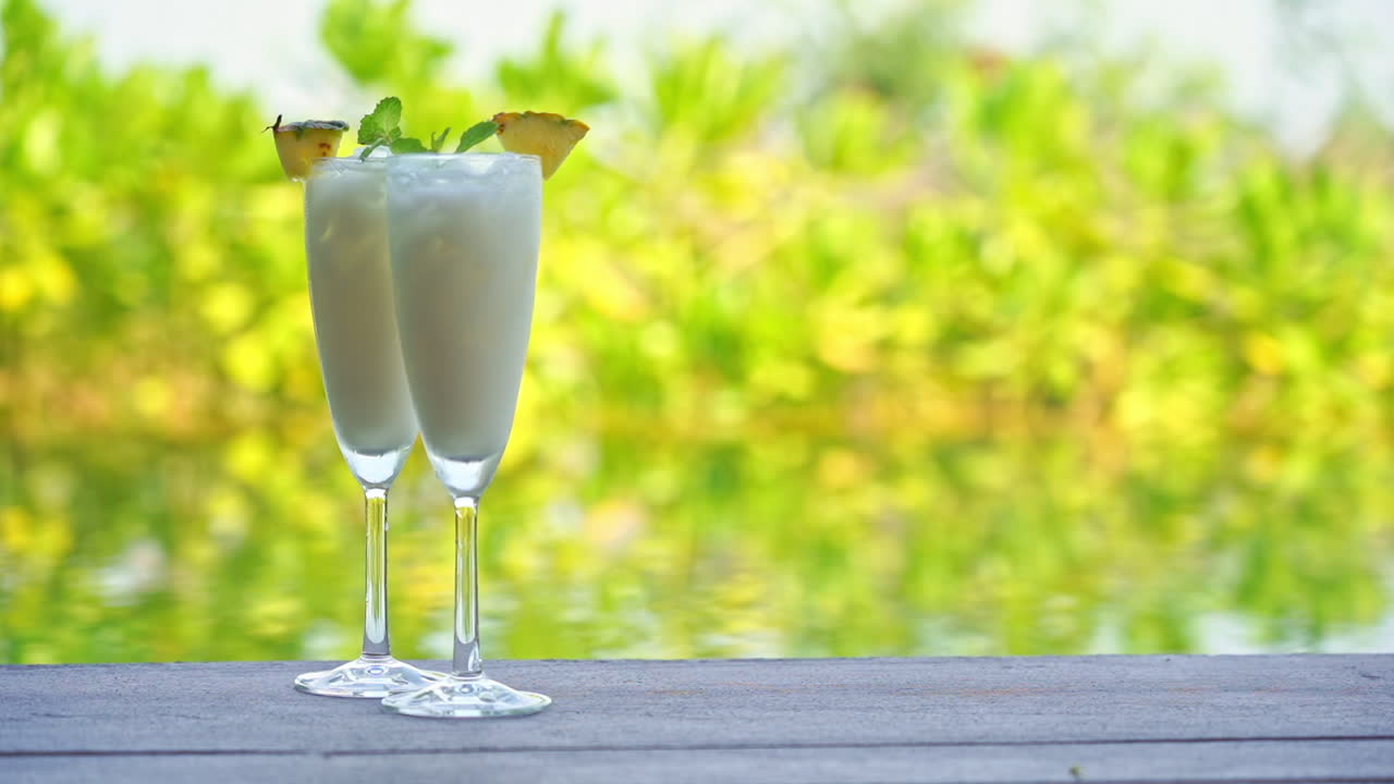 Two tall glasses filled with milky coconut frozen drink and pineapple garnish on wooden table with shimmering water and tropical green foliage bokeh background. Refreshing tropical drink