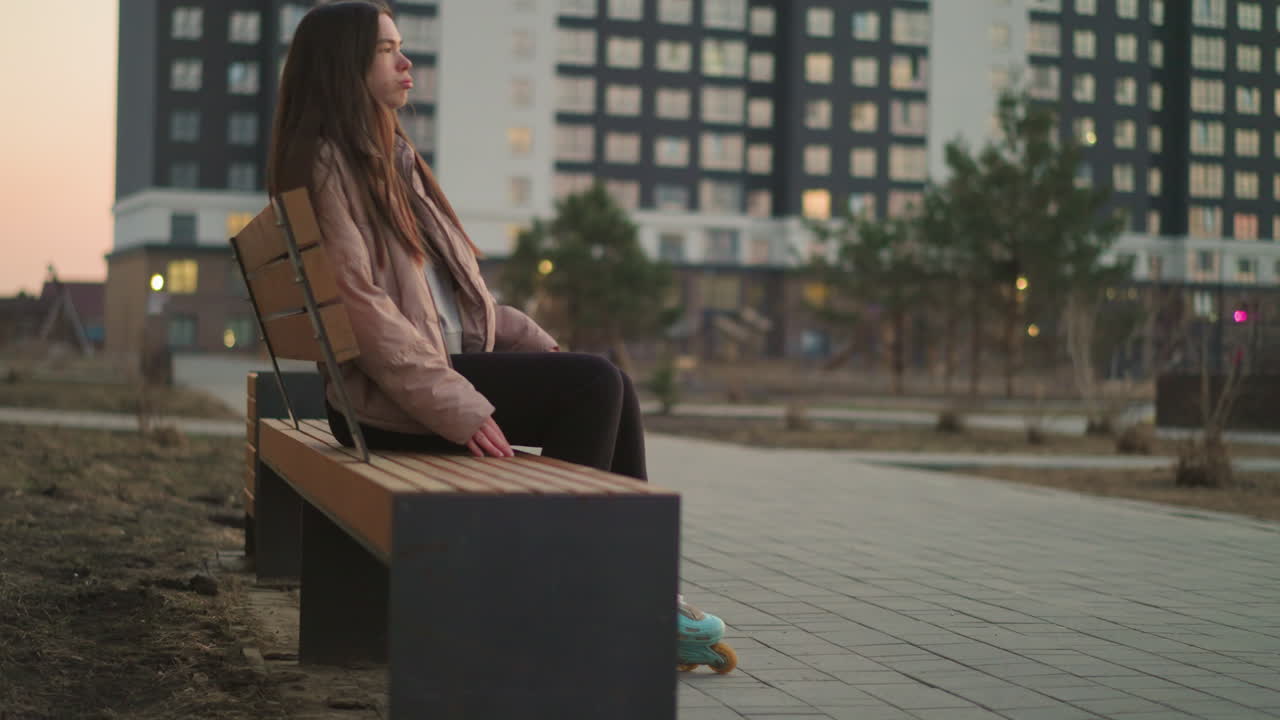 A girl in a peach jacket and black trousers is seen rollerblading towards a park bench. The image captures the moment before she sits down to rest, with her rollerblades visible on the paved path