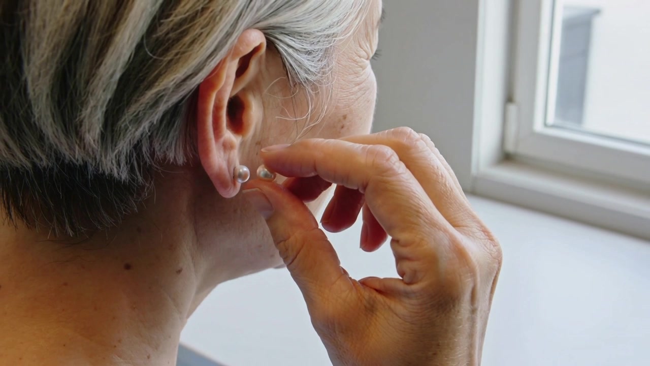 Femme asiatique âgée ramassant la deuxième boucle d'oreille en perle, la mettant au comptoir près de la fenêtre, téléphone visible.