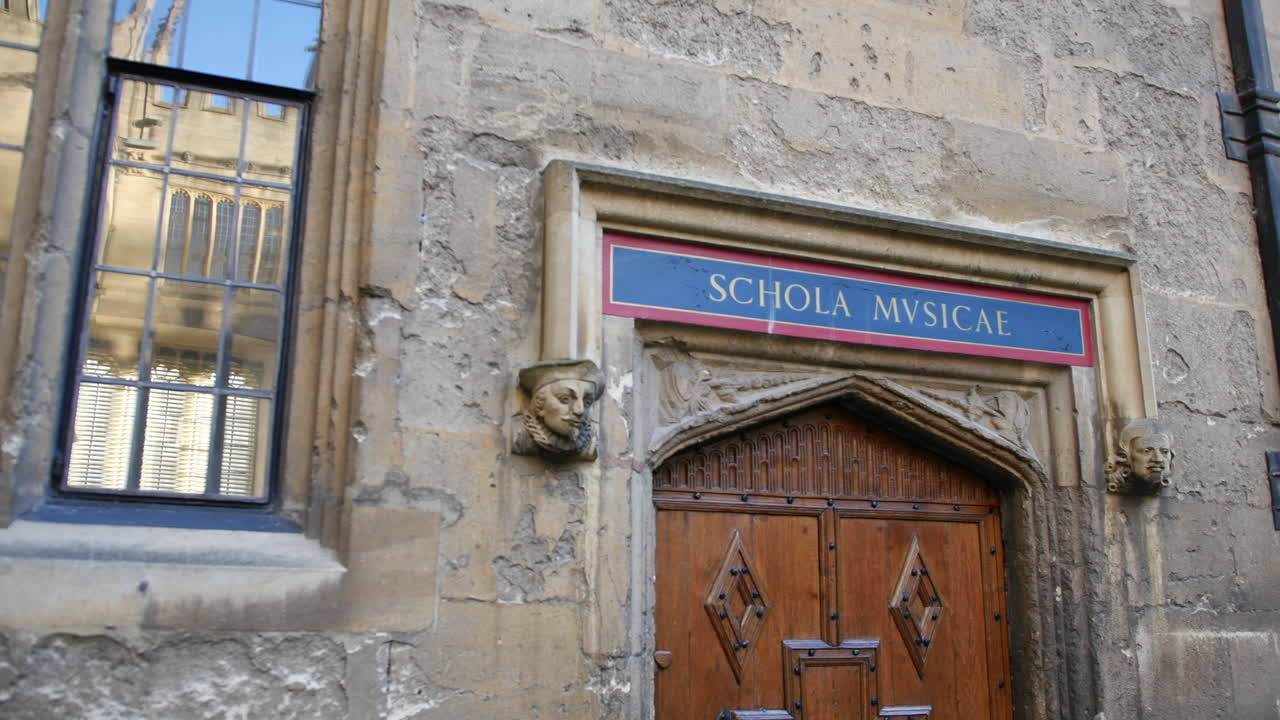 Detalhe de portal na Universidade de Oxford mostrando Schola Musicae, que significa escola de música, com rostos esculpidos em pedra, texturas de parede envelhecidas e janela de vitral em close-up